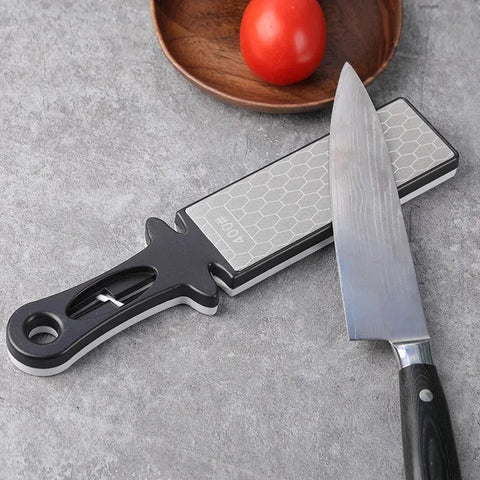 Knife sharpener and a chef's knife on a gray surface with a wooden bowl and tomato in the background.