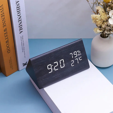 Digital clock displaying time, temperature, and humidity on a blue surface with books and a vase in the background.