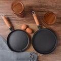 Two black frying pans with wooden handles on a wooden surface with eggs and jars.