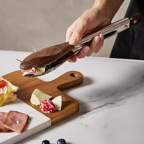 Hand using tongs to pick up a sandwich from a wooden cutting board on a marble surface.