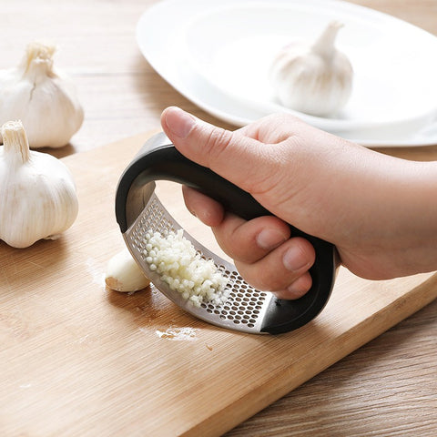 Hand using a garlic press on a wooden cutting board with garlic cloves and a white plate in the background.