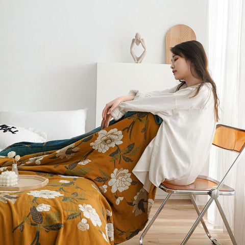 Woman sitting on a chair with a floral blanket draped over her, in a room with a bed and decor.