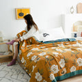 Cozy bedroom scene featuring a golden yellow cotton blanket with white floral patterns and a teal border draped over a bed, styled with soft pillows and natural light filling the room.
