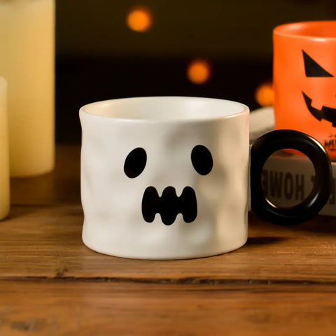 White mug with ghost face design on a wooden surface with Halloween-themed items in the background.