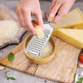 Person using a handheld grater on a piece of cheese with a wooden cutting board and ingredients in the background.
