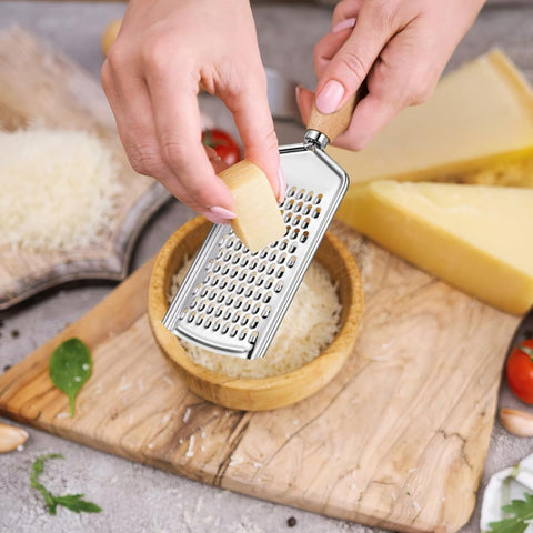 Person using a handheld grater on a piece of cheese with a wooden cutting board and ingredients in the background.