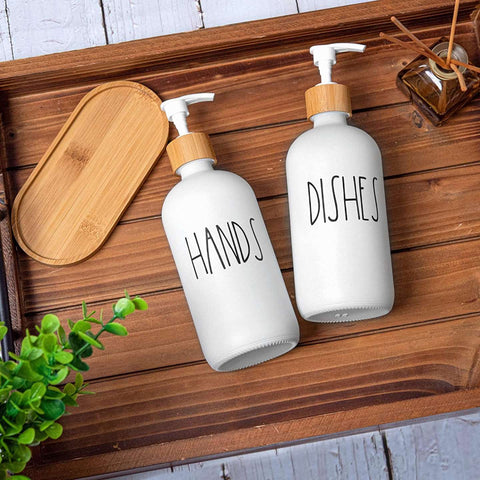 Two white bottles labeled 'HANDS' and 'DISHES' on a wooden surface with a wooden tray and plant in the background.