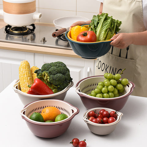 Set of multi-colored colanders with fresh fruits and vegetables on a kitchen counter.