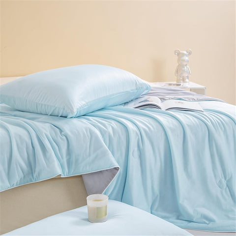 Soft light blue ice silk quilt with matching pillow on a cozy bed, styled with a book and candle in a warm bedroom setting.