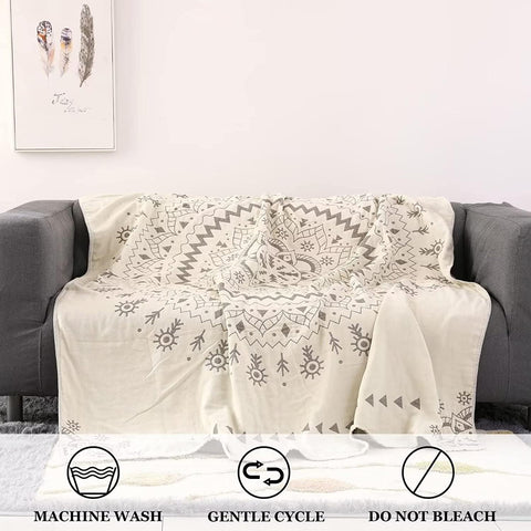  Beige cotton boho throw blanket with gray tribal patterns draped over a gray sofa, showing care icons for machine wash, gentle cycle, and no bleach.