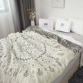 Beige cotton boho throw blanket with gray tribal patterns and contrasting gray border, softly draped over a bed in a bright room with decorative bags and dried flowers in the background.