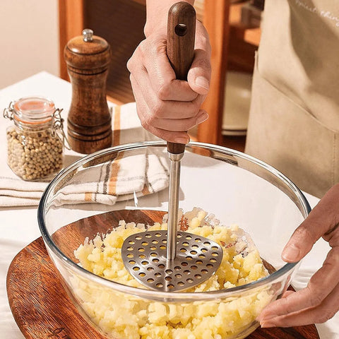 Person mashing potatoes in a glass bowl with a metal masher, kitchen setting.