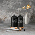 Black 'HANDS' and 'DISHES' bottles with a white vase with flowers on a textured gray background