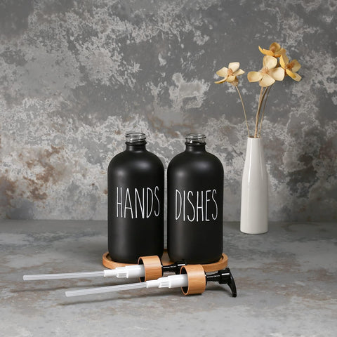 Black 'HANDS' and 'DISHES' bottles with a white vase with flowers on a textured gray background