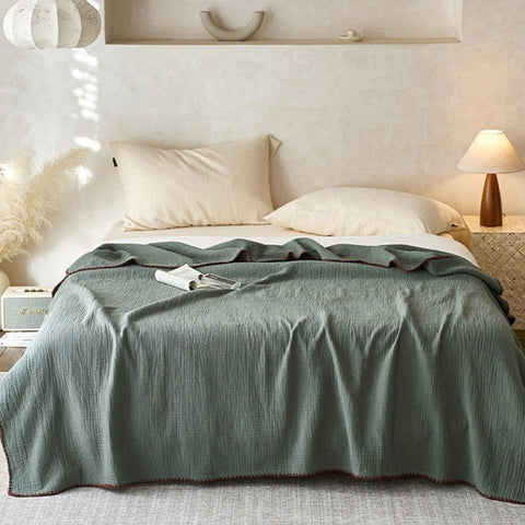 Olive green cotton muslin blanket with dark trim styled on cozy bed