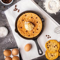 Skillet cookie with ice cream surrounded by baking ingredients on a dark surface