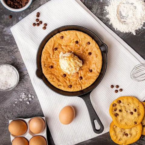 Skillet cookie with ice cream surrounded by baking ingredients on a dark surface