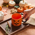 Orange mug with Halloween design held by a hand, surrounded by pumpkins and autumn leaves on a wooden tray.