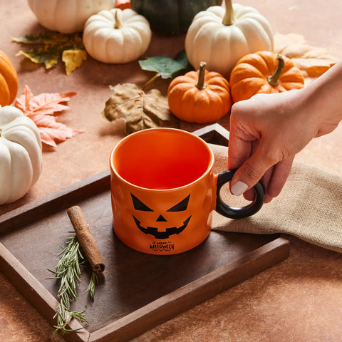 Orange mug with Halloween design held by a hand, surrounded by pumpkins and autumn leaves on a wooden tray.