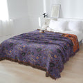 Purple boho cotton throw blanket with floral pattern draped on a bed in a bright, minimalist bedroom setting with white pillows and soft natural light