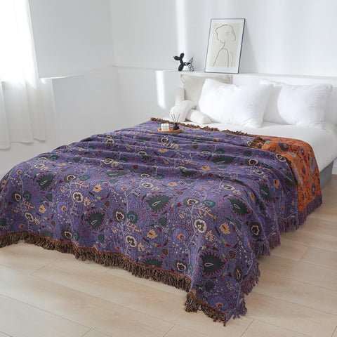 Purple boho cotton throw blanket with floral pattern draped on a bed in a bright, minimalist bedroom setting with white pillows and soft natural light