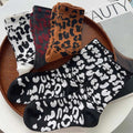Assorted leopard print socks in black, white, brown, and gray colorways displayed on a wooden tray, featuring bold retro patterns and soft ribbed fabric.