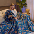 Woman sitting on a bed wrapped in a colorful floral blanket in a cozy room holding a cup while smiling. 