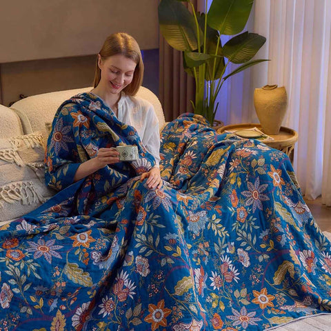 Woman sitting on a bed wrapped in a colorful floral blanket in a cozy room holding a cup while smiling. 