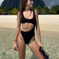 Black ruched one-piece swimsuit with front cutout shown on model at the beach.