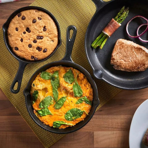 Three small cast iron skillets with different food items on a wooden surface.