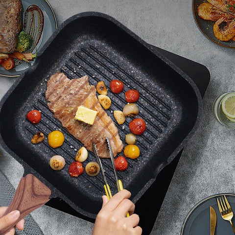 Person grilling steak and vegetables on a black grill pan with a wooden spatula.