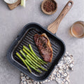 Grilled steak and asparagus in a black grill pan on a gray surface with a glass of beer and wooden spoon.