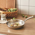 Stainless steel frying pan on a wooden surface with vegetables and a basket in the background