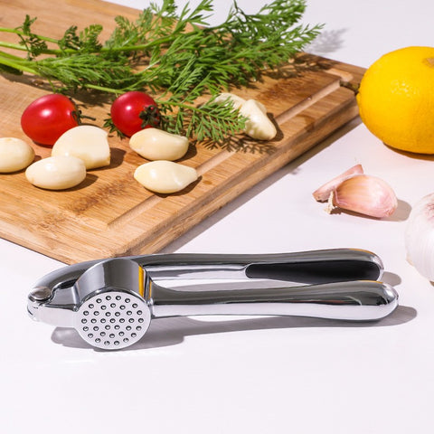 Garlic press on a white surface with vegetables and a lemon in the background
