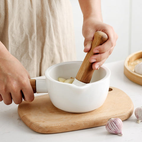 Hands holding white ceramic mortar and wooden pestle crushing garlic cloves on wooden cutting board with garlic bulbs nearby