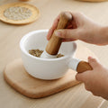 Hands holding white ceramic mortar with wooden handle and pestle crushing spices on wooden board
