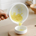 Hand pouring crushed garlic from white ceramic mortar with wooden handle into small matching bowl on wooden surface