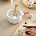 White ceramic mortar and pestle with wooden handle on table next to hands cutting fresh garlic on wooden board