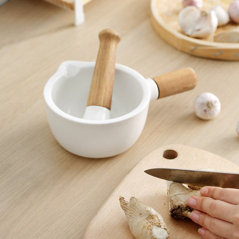 White ceramic mortar and pestle with wooden handle on table next to hands cutting fresh garlic on wooden board