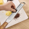 Hand grating chocolate on a wooden handle stainless steel grater over white plate with grated chocolate and cheese on wooden table