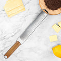 Wooden handle stainless steel grater placed on marble countertop with slices of cheese, lemon, and grated chocolate in wooden bowl nearby
