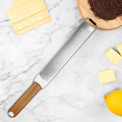Wooden handle stainless steel grater placed on marble countertop with slices of cheese, lemon, and grated chocolate in wooden bowl nearby