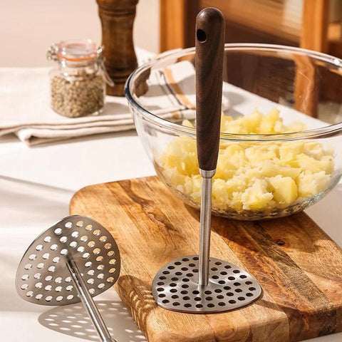 Clear glass bowl with wooden-handled masher on a wooden cutting board with mashed potatoes.