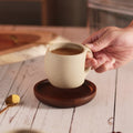 A beige ceramic coffee mug with a gold-colored spoon in it, resting on a brown wooden saucer, which is placed on a wooden surface, hand reaching for a mug.